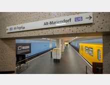 U-Bahnhof mit blauen Kacheln an den Wänden, einem gelben Zug der Linie U6 auf der rechten Seite und einem Hinweisschild mit den Stationen „Alt-Tegel“ und „Alt-Mariendorf“. Im Vordergrund befinden sich eine Karte und ein Werbeplakat.