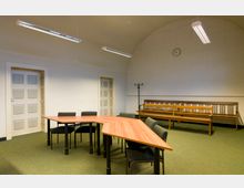 Ein schlichter Besprechungsraum mit einem dreieckig angeordneten Holztisch und mehreren gepolsterten Stühlen auf grünem Teppichboden. Im Hintergrund befinden sich zwei Holzbänke, eine Wanduhr, eine geschwungene Decke und zwei Türen mit Kassettenstruktur.