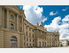 Außenansicht eines historischen, barocken Gebäudes mit geschwungener Fassade, Säulen, großen Fenstern und zahlreichen Statuen auf dem Dach. Der Himmel ist blau mit einigen Wolken, und im Vordergrund befindet sich ein gepflasterter Platz.