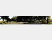 Panoramaaufnahme einer Landstraße, die von Bäumen gesäumt ist und eine leichte Kurve macht. Auf der rechten Seite befindet sich ein kleines Gebäude mit Wegweisern und ein Leitplanke zieht sich entlang der Straße.