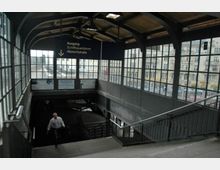 Bahnhofseingang mit überdachtem Treppenbereich und großen, rechteckigen Fensterfronten, die den Blick auf umliegende Gebäude und Bahngleise freigeben. Ein Schild weist auf den Ausgang "Schiffbauerdamm/Albrechtstraße" hin.