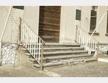 Eingangsbereich eines Gebäudes mit mehreren abgenutzten Steinstufen und einem Metallgeländer. Die Holztür und Fenster mit geschlossenen Rollläden sind in eine schlichte Fassade aus Putz und Stein eingelassen.