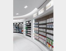 Innenraum einer modernen Apotheke mit einer abgerundeten weißen Wand und Regalen, die Hautpflege- und Kosmetikprodukte präsentieren. Oben sind beleuchtete Schriftzüge mit den Worten "Kosmetik", "Körper" und "Haut" zu sehen, während die Regale Produkte in weißen und farbigen Verpackungen ordentlich ausgestellt zeigen.