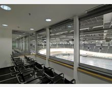 Blick aus einer verglasten Zuschauerloge auf eine große Indoor-Sporthalle mit einer Radrennbahn und Sitztribünen im Hintergrund. Im Vordergrund befinden sich mehrere schwarze Stühle mit Metallrahmen auf Teppichboden.