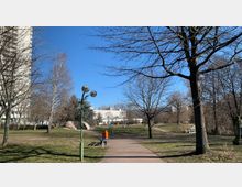 Ein städtischer Park mit gepflastertem Gehweg, umgeben von kahlen Bäumen und Grasflächen. Im Hintergrund sind moderne Gebäude zu sehen, während mittig eine Straßenlaterne und eine Bank platziert sind, rechts vom Weg steht ein orangefarbener Abfalleimer.