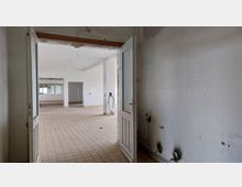 Blick in einen verlassenen Raum mit offen stehender Holztür. Der Raum ist gefliest, hat helle Wände mit Gebrauchsspuren und sichtbaren Bohrstellen, während Leitungen lose heraushängen.