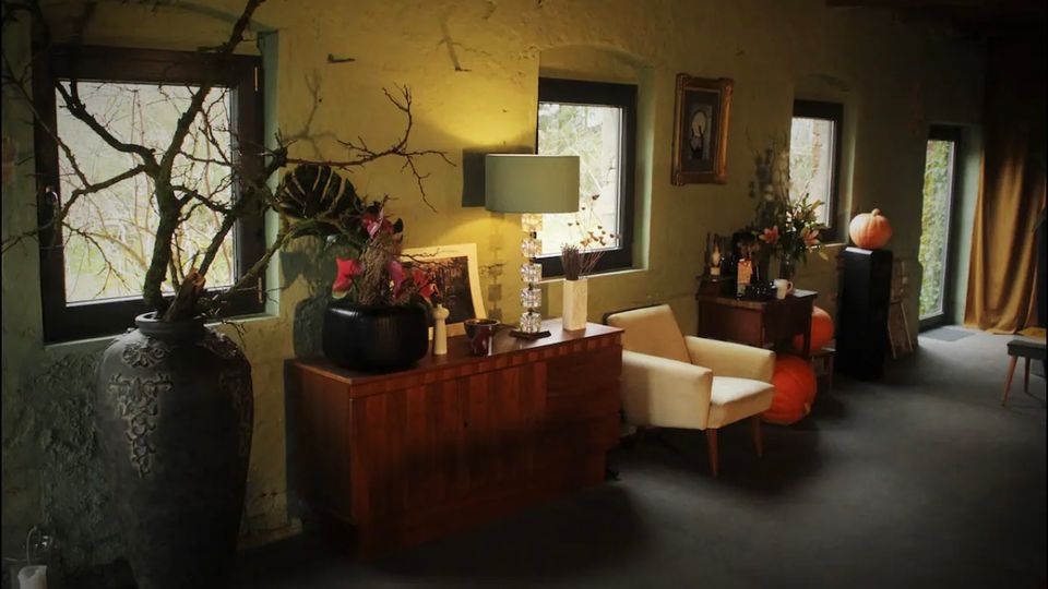 Wohnraum mit rustikalem Stil, grün gestrichenen Wänden und mehreren Fenstern, die natürliches Licht hereinlassen. Der Raum ist dekoriert mit großen Vasen, einer Kommode mit Lampe und Blumenarrangements, sowie einem Sessel und herbstlichen Akzenten wie Kürbissen.