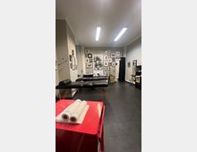 Ein heller Tätowierladen mit einem schwarzen Massagetisch in der Mitte, dekoriert mit einer Wand voller gerahmter Kunstwerke und Zeichnungen. Im Vordergrund steht ein roter Wagen mit aufgerolltem Papier, der Boden ist mit dunklen Fliesen ausgelegt.