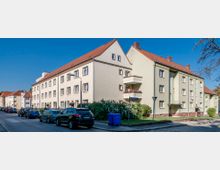 Mehrfamilienhäuser im Stil der Nachkriegszeit mit beigefarbenen Fassaden und roten Satteldächern an einer Straßenecke. Parkende Autos stehen entlang der Straße, und im Vordergrund ist ein blauer Müllcontainer zu sehen.