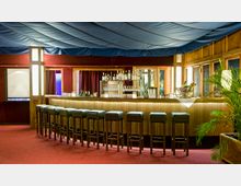 Eine elegante Bar mit gepolsterten Hockern aus dunklem Leder, einer langen, hölzernen Theke und einer Rückwand mit beleuchteten Regalen, die Flaschen und Gläser präsentieren. Der Raum hat rote Teppichböden, eine Stoffdecke in Blau und Pflanzen als Dekoration.