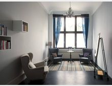 Ein heller Raum mit einem großen Fenster, blauen Vorhängen und einem Kronleuchter an der Decke. Zwei graue Sessel stehen auf einem gestreiften Teppich, dazwischen ein kleiner runder Tisch; an der Wand sind Regale mit Büchern angebracht, und im Vordergrund steht ein Flipchart.