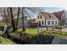 Ein kleines ländliches Wohnhaus mit heller Fassade und rotem Ziegeldach steht neben weiteren Häusern in einer Dorfumgebung. Im Vordergrund befinden sich ein schmaler Bachlauf, Rasenflächen und mehrere Bäume, die einen Zaun um das Anwesen säumen.