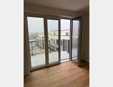 Blick aus einem modernen Wohnraum mit Holzfußboden auf eine große Terrassentür, die zur Hälfte geöffnet ist. Die Terrasse hat Fliesenboden und ein Metallgeländer; im Hintergrund sind beige Wohngebäude und eine verschneite Umgebung zu sehen.