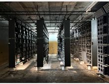 Ein großer Raum mit mehreren Reihen von Metallregalen, die mit elektrischen Bauteilen und Verkabelungen bestückt sind. Die Beschriftung auf den Metallstützen zeigt Nummerierungen (z. B. "Gest. 21-28"), und an der Decke verlaufen Kabel und Rohrleitungen.