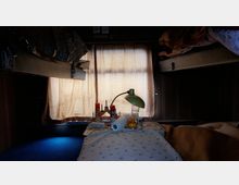 Innenraum eines Schlafabteils in einem Zug oder Wohnbereich mit zwei Etagenbetten an den Seiten, einem kleinen Tisch in der Mitte mit einer Lampe, Lebensmitteln und Papierrolle darauf. Im Hintergrund sind geschlossene Vorhänge zu sehen, die das Licht dimmen.