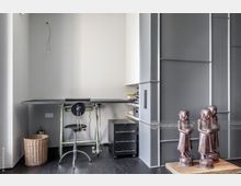 Minimalistisch eingerichtetes Büro mit einem schwarzen höhenverstellbaren Zeichnungstisch und schwarzem Drehstuhl vor einer grauen Wand. Rechts befinden sich graue Einbauschränke, darunter ein Regal mit Büchern und ein Drucker; im Vordergrund stehen drei dekorative Holzskulpturen auf einem Holzpodest.