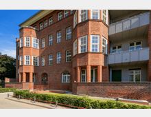 Rotes Backsteingebäude mit mehreren Etagen, markanten Erkern mit weißen Sprossenfenstern und einem Eingangsbereich mit Rundbogen; davor eine kleine Grünfläche mit Hecken.