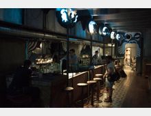 Dunkle Innenaufnahme einer Bar mit langem Tresen und hohen Barhockern. Die Bar ist in einem industriellen Stil gestaltet, beleuchtet von dekorativen, blauen Lampen, mit Wandmustern und Personen, die an der Bar sitzen oder arbeiten.