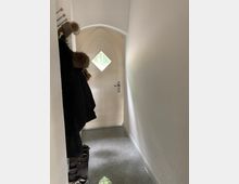 Schmaler Flur mit grauem Fliesenboden, links eine Garderobe mit Jacken und Mützen, rechts weiße Wände, am Ende eine cremefarbene, spitzbogige Tür mit kleinem rautenförmigem Fenster.