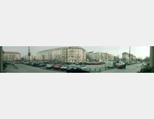 Panorama einer städtischen Platzszene mit parkenden Autos in der Mitte, flankiert von mehrstöckigen Wohn- und Geschäftshäusern im Hintergrund. Die Straße und der Platz sind mit Pflastersteinen bedeckt, und vereinzelt stehen Laternen sowie kleine Bäume.