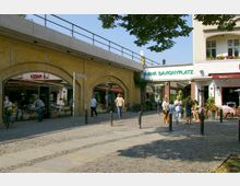 Straßenszene am S-Bahnhof Savignyplatz in Berlin mit Geschäften in gelben Klinkerbögen, Passanten und einem kleinen Platz mit Pflastersteinen; im Hintergrund ist ein Schild mit der Aufschrift „S-Bhf. Savignyplatz“ zu sehen.
