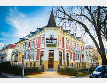 Eckgebäude im Stil der Gründerzeit mit spitzem Turmdach, roter Klinkerfassade und hellen Fenstereinfassungen. Vor dem Gebäude befinden sich ein schmiedeeiserner Zaun und ein Straßenschild mit der Aufschrift "Jägerstraße", daneben ein Baum ohne Laub.