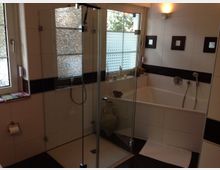 Modernes Badezimmer mit weiß gefliesten Wänden und dunklen Akzenten. Es umfasst eine Glasdusche, eine Badewanne, ein Fenster mit Milchglas und Sichtschutz sowie dezente Beleuchtung.