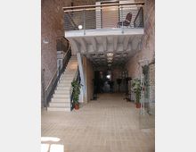 Innenansicht eines modernen Gebäudes mit Backsteinwänden, einer Treppe aus Beton und Stahlgeländer auf der linken Seite, und einem offenen Zwischengeschoss mit Glasbrüstung. Der Boden ist mit hellen Fliesen bedeckt, und Pflanzen in Töpfen lockern den Raum auf.