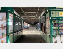 Unter einer Überführung befinden sich grüne Kioske mit teilweise heruntergelassenen Rollläden und Plakaten, die Speisen und Getränke bewerben. Im Hintergrund sind Fahrradständer mit abgestellten Fahrrädern und ein überdachter Gehweg erkennbar.