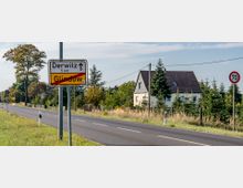 Landstraße mit Verkehrszeichen, das das Ende des Ortes Glindow und die Richtung nach Derwitz (5 km) anzeigt. Im Hintergrund ein Wohnhaus mit Garten, umgeben von Bäumen und Wiesen, sowie ein Schild für die Geschwindigkeitsbegrenzung von 70 km/h.