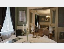 Eleganter Innenraum mit klassischer Einrichtung: Ein runder Tisch mit Tischdecke und einem Kerzenhalter im Vordergrund, eine geöffnete Doppeltür führt zu einem angrenzenden Raum mit Fenstern, Vorhängen, Gemälden und einem Kronleuchter an der Decke. Die Wände sind aufwendig mit grünen Tapeten und Zierelementen gestaltet.