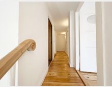 Langer, heller Flur in einem Wohngebäude mit Holzdielenboden, weißen Wänden und Türen. Links ist ein glatt geschliffenes Holzgeländer mit eingerolltem Endstück sichtbar.