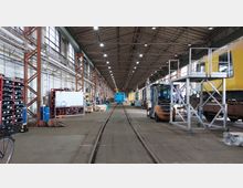 Innenansicht einer großen Werkhalle mit Schienen, die zentral durch den Raum verlaufen. Links stehen Regale mit Arbeitsmaterialien und ein Whiteboard, während rechts ein Hebe-Gerüst und ein gelber Zug sichtbar sind. Am Ende der Halle befindet sich ein blauer Zug.