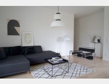 Modernes Wohnzimmer mit grauem Ecksofa, schwarzem Couchtisch und einem gemusterten Teppich. An der Wand hängen minimalistische Kunstwerke, daneben stehen eine weiße Stehlampe und ein schwarzer Designersessel auf hellem Holzboden.