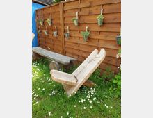 Holzzaun aus braunen Latten mit daran befestigten Pflanztöpfen aus Metall, die kleine Pflanzen und Kräuter enthalten. Davor stehen eine rustikale Holzbank und ein ebenfalls rustikaler Holzstuhl auf einer Wiese mit blühenden Gänseblümchen.