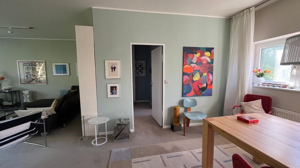 Heller Wohn- und Essbereich mit hellgrünen Wänden, großem Fenster mit Vorhang und Teppichboden; an den Wänden hängen gerahmte Bilder und ein großes farbiges Gemälde. Rechts steht ein Holztisch mit roten Stühlen, daneben ein Sitzbereich mit Sofa, Beistelltisch und weiteren Stühlen sowie eine offene Tür zum Flur.