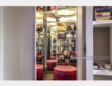 Ein begehbarer Kleiderschrank mit gläsernen Regalen, die mit Designer-Handtaschen gefüllt sind. Der Raum ist mit Spiegelwänden ausgestattet, und in der Mitte befindet sich ein runder, pinkfarbener Sitzhocker auf einem dunklen Holzboden.