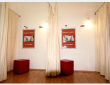 Ein Umkleidebereich mit hellen Vorhängen, Holzboden und zwei roten Sitzhockern. An der weißen Wand hängen Poster mit rotem Hintergrund, und die Szene wird von modernen Wandlampen beleuchtet.