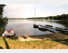 Seeufer mit kleinem Steg und mehreren festgemachten Booten, umgeben von Wiesen und Bäumen. Im Hintergrund ist ein ruhiger See mit bewaldeter Uferlinie zu sehen.