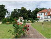 Weißes Wohnhaus mit roten Ziegeldächern und grünen Fensterläden, umgeben von einer gepflegten Rasenfläche und Rosenbüschen; im Hintergrund Bäume und weitere Gebäude. Ein gepflasterter Weg mit einer Laterne führt durch die Grünanlage.