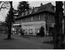 Schwarz-weiß-Foto eines zweistöckigen Gebäudes mit Satteldach, Schaufenstern im Erdgeschoss und mehreren kleinen Fenstern im Dachgeschoss. Im Vordergrund sind Bäume, ein gepflasterter Platz und ein Eingang zu einem Geschäft oder Servicebereich sichtbar.