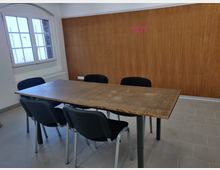 Ein einfacher Besprechungsraum mit einem Holztisch, umgeben von vier schwarzen Stühlen. Im Hintergrund befinden sich eine weiße Wand mit einem Fenster und eine Holzverkleidung mit der Aufschrift „TRY!“ in pinker Schrift.