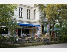 Eckcafé mit Außenbereich, umgeben von Bäumen und verziert mit einem schmiedeeisernen Zaun; blaue Markise mit dem Schriftzug „Yorckschlösschen“ und bunte Lichterketten. Im Vordergrund steht ein Fahrrad an einem Baum, im Hintergrund sind Tische und Sonnenschirme zu sehen.