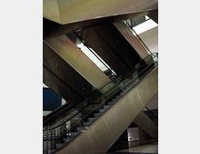 Ein Innenraum mit modernen Rolltreppen, die sich diagonal über mehrere Etagen erstrecken. Die Struktur besteht aus Metall und Glas, mit einer minimalistisch gestalteten Umgebung und hellen Decken.