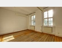 Leerer Raum mit weiß gestrichenen Wänden, einem Holzdielenboden und großen Sprossenfenstern, durch die Tageslicht einfällt. Unter den Fenstern befinden sich Heizkörper, und an der Decke hängt eine einfache Leuchtstofflampe.