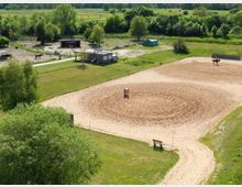 Luftaufnahme eines Reitplatzes mit sandigem Boden, auf dem zwei Pferde stehen. Im Hintergrund befinden sich grüne Wiesen, Bäume, mehrere Unterstände sowie ein kleines Gebäude.