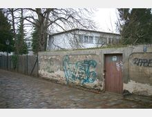 Gepflasterte Gasse entlang einer alten, mit Graffiti besprühten Steinmauer, die eine Holztür enthält; dahinter ist ein flaches, modernes weißes Gebäude und kahles Baumwerk sichtbar.