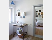 Ein schlichtes Badezimmer mit weiß gestrichenen Wänden und einem kleinen Fenster auf der linken Seite. Ein Waschbecken auf einem rustikalen Metall- und Holzgestell wird von einem Spiegel mit Holzrahmen darüber ergänzt; darunter stehen Handtücher in einem Korb. Eine offene Tür führt in ein Schlafzimmer mit Holzfußboden und einer dekorierten Wand mit Bildern.