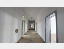 Langer, schmaler Flur mit weißen Wänden und einer Reihe von Türen auf beiden Seiten, einige mit Oberlichtern versehen. Der Boden ist roh und unbehandelt, und ein Loch in der Wand auf der linken Seite ist sichtbar.