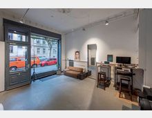 Ein heller, offener Raum mit hohen Decken, großen Schaufenstern und Blick auf eine Straße mit parkenden Autos. Der Raum ist mit einem Sofa, einem Schreibtisch mit Computern, einem großen Spiegel und weiteren minimalistischen Möbeln ausgestattet.